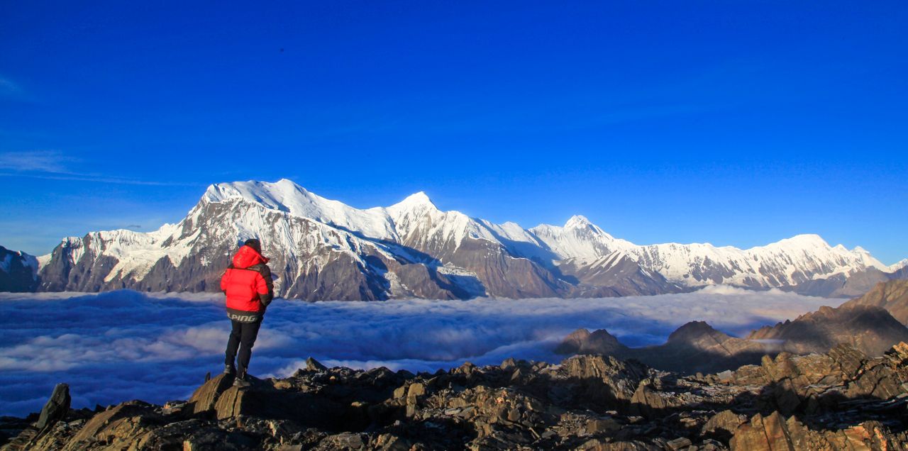 Back view of anonymous male traveler in outerwear standing on rocky surface while contemplating mountains with snow on peaks under bright cloudless sky in wintertime